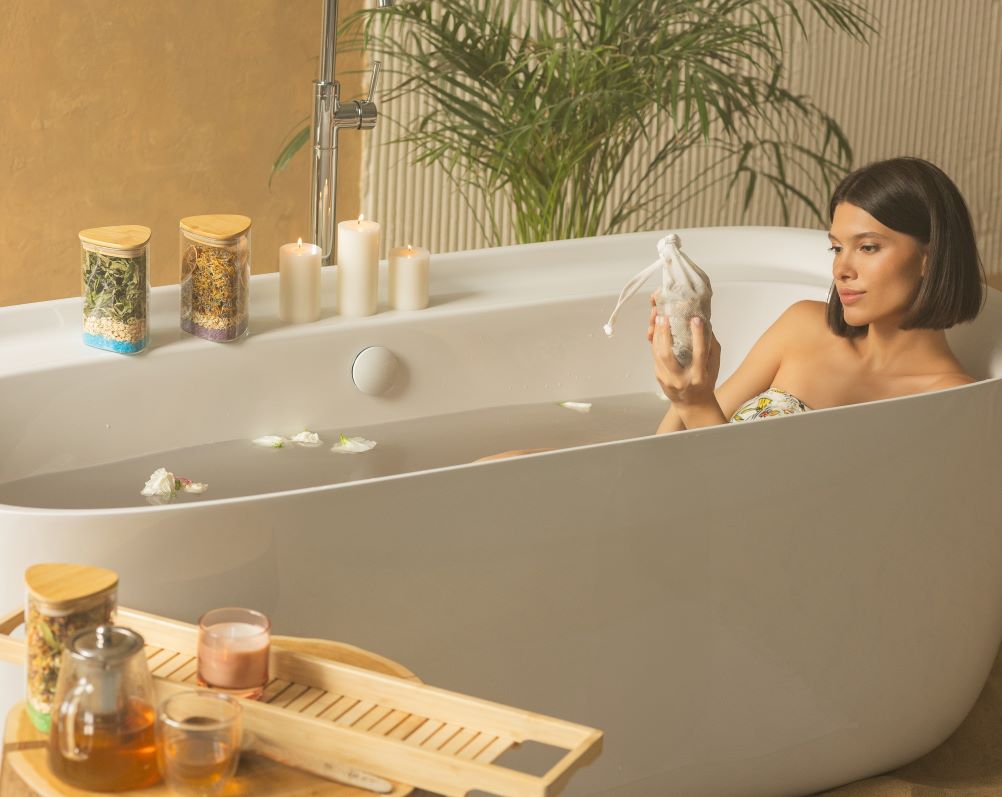 A woman enjoys a tranquil bath, surrounded by candles, with Bath Tea Blend soothing and squeezing a tea bag with plants for aromatherapy.