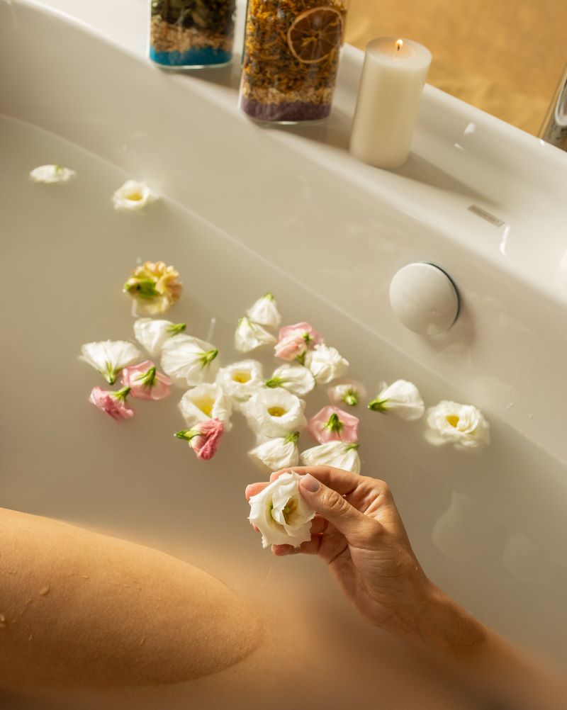 A woman enjoys a serene bath surrounded by vibrant flowers, enhancing her relaxation with a soothing blend of plants and essential oils.